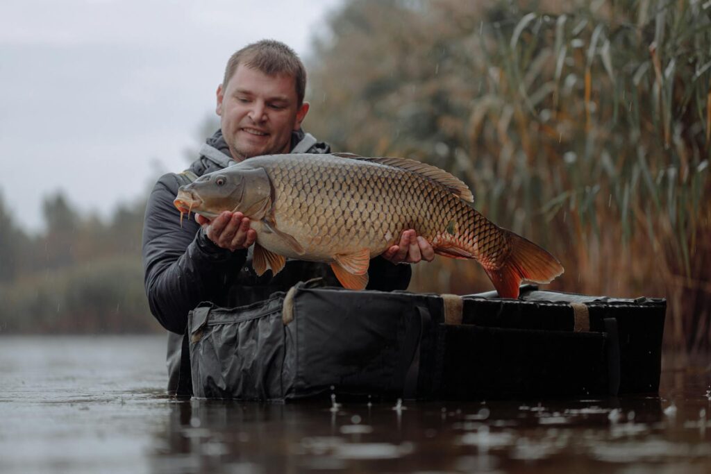 A fisherman proudly displays his catch, a large carp, by a serene, rainy lake.